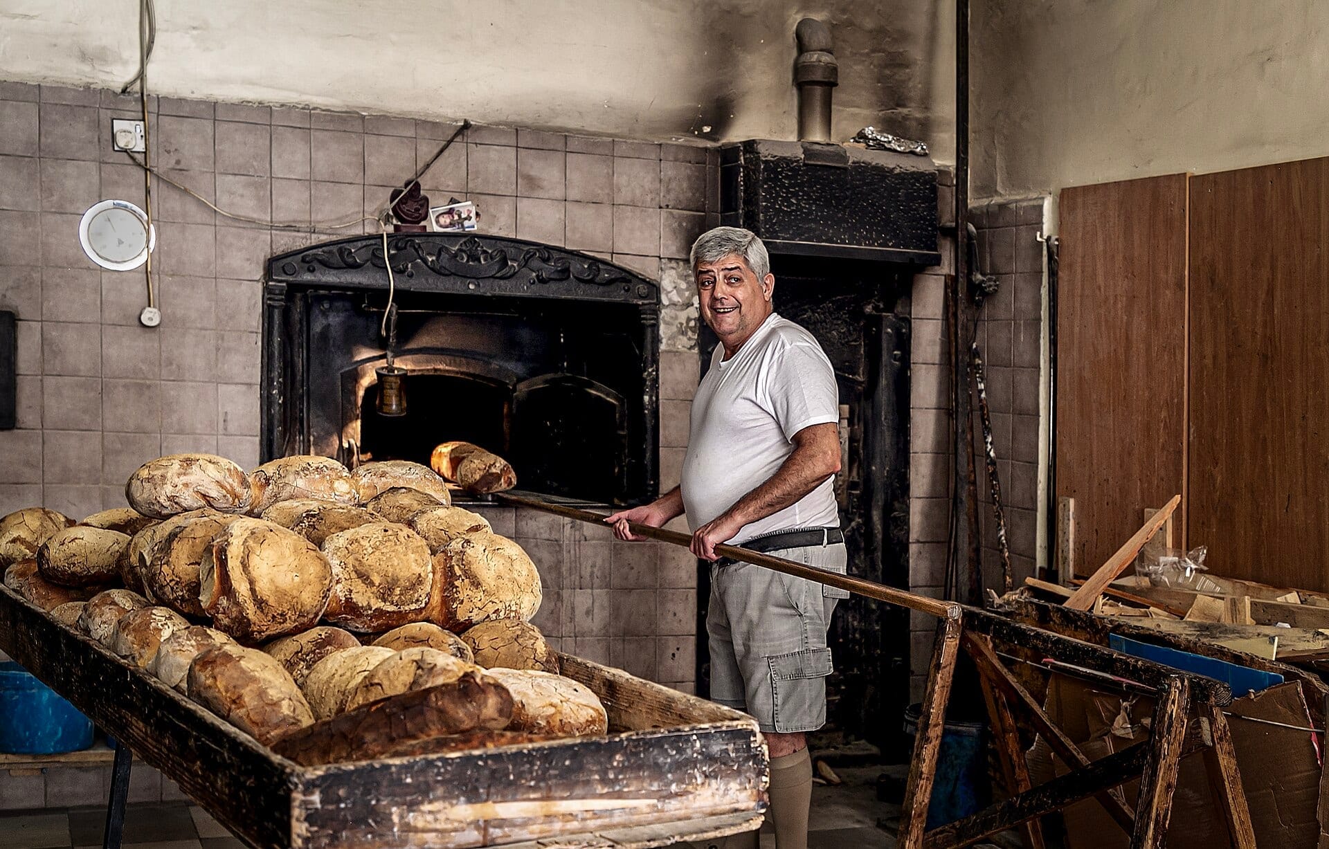 A baker with a long wooden peel slides loaves into a brick oven beside a rack of rustic round loaves