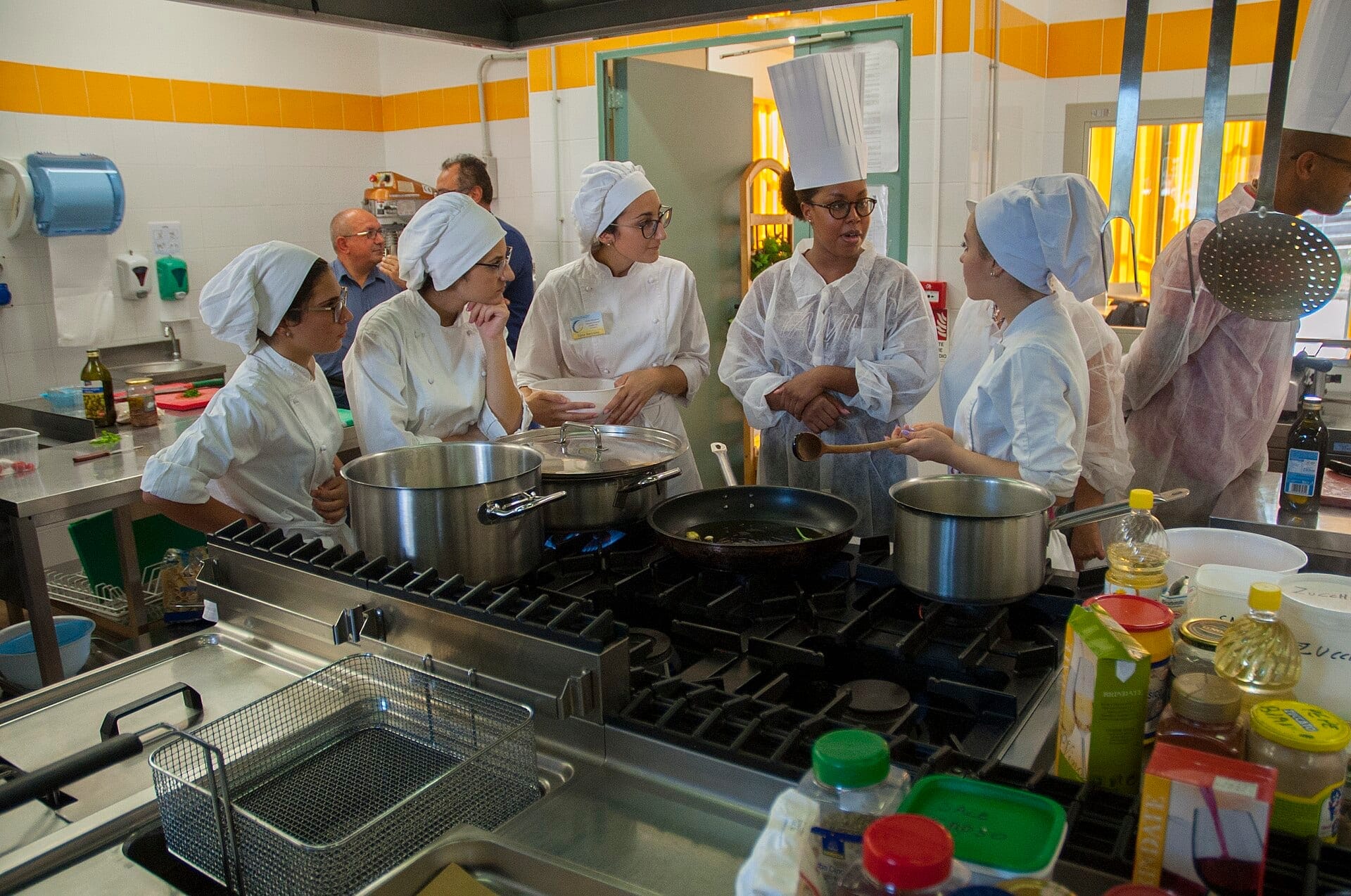 Culinary students in white jackets and caps gather around a gas range in a tiled teaching kitchen while an instructor speaks