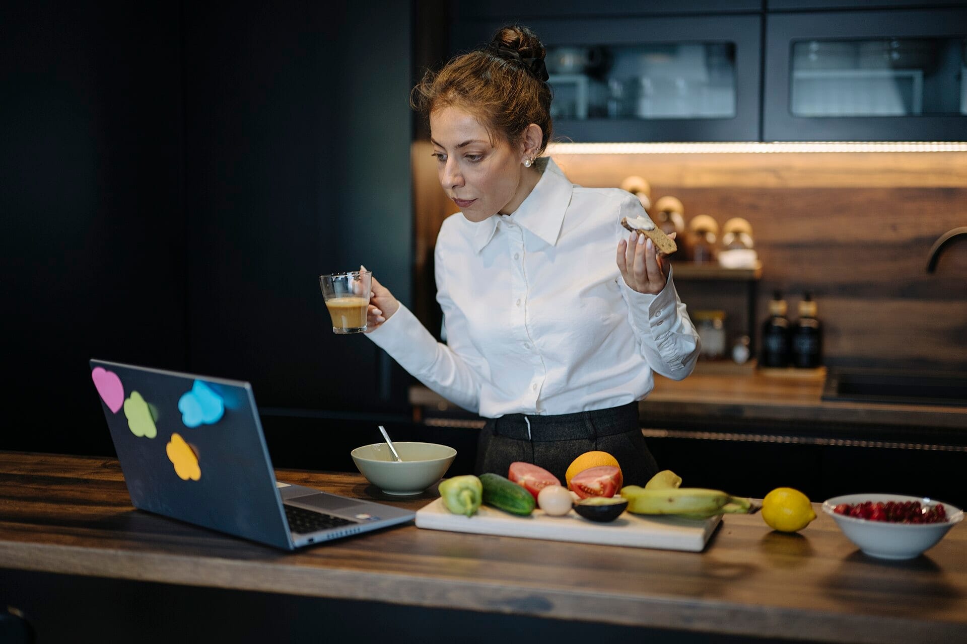 A woman in a white shirt looks at an open laptop in a dark kitchen with chopped vegetables and fruit on the counter