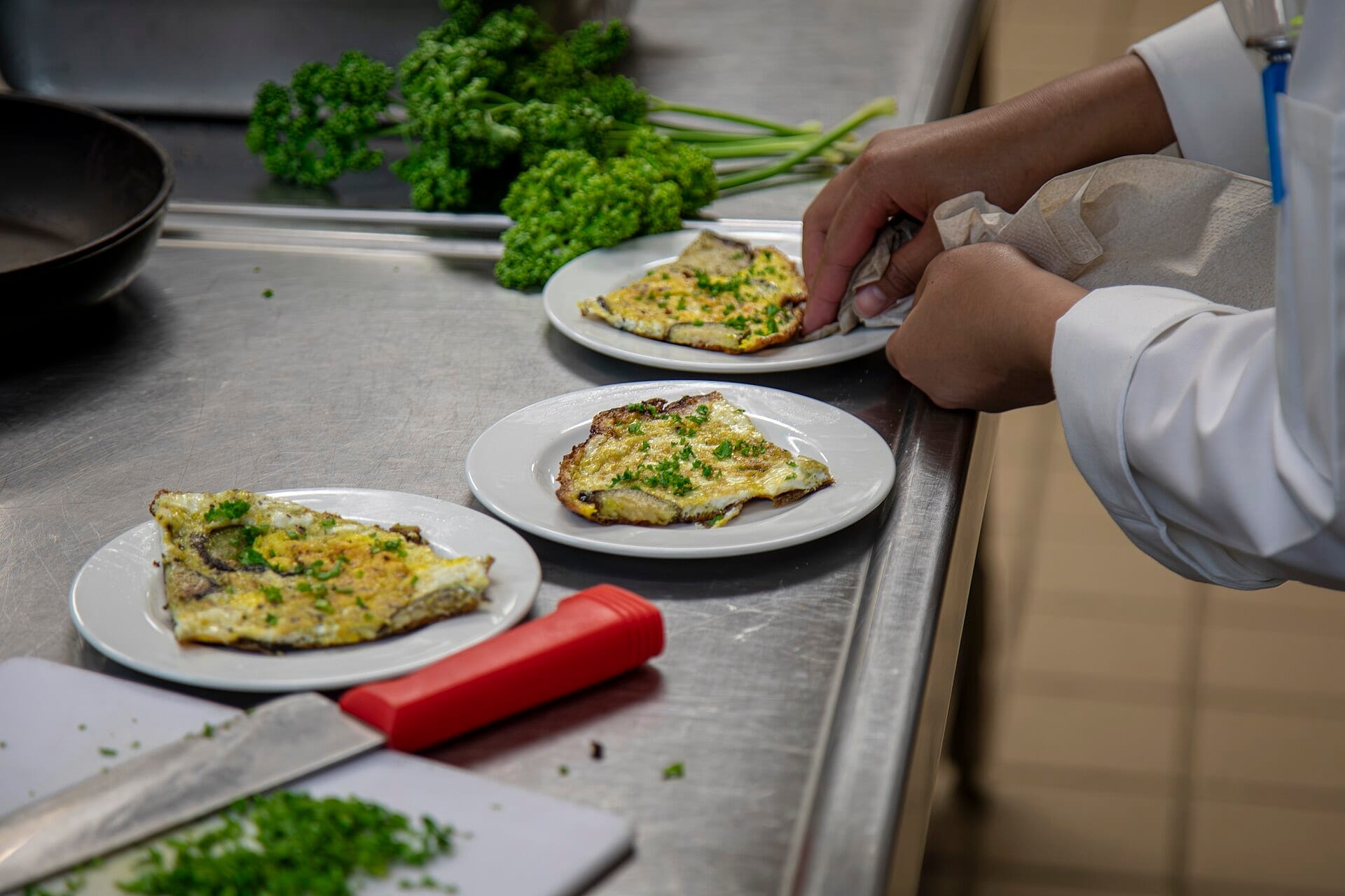 A cook's hands in a white jacket wipe the rim of a plate of herb omelette on a stainless steel pass, with a knife and chopped parsley nearby
