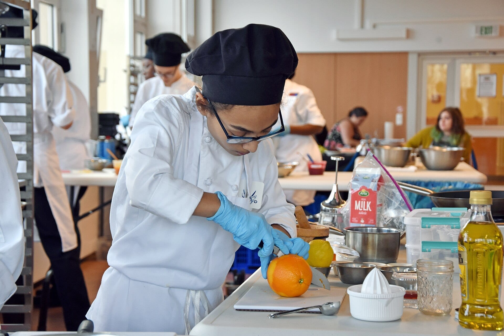 A student in chef whites and a black cap cuts an orange on a board in a bright shared teaching kitchen