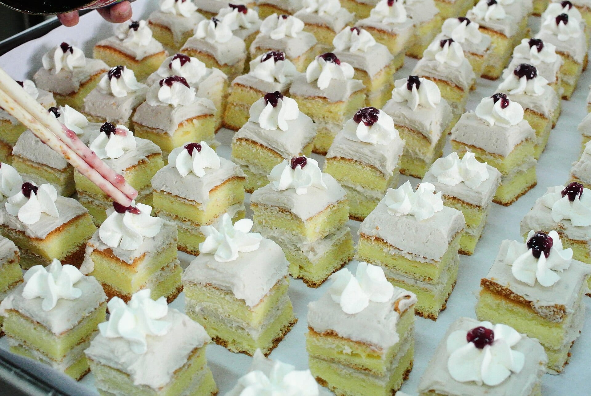 Rows of small sponge cakes topped with cream rosettes and a berry, being finished with chopsticks on a tray
