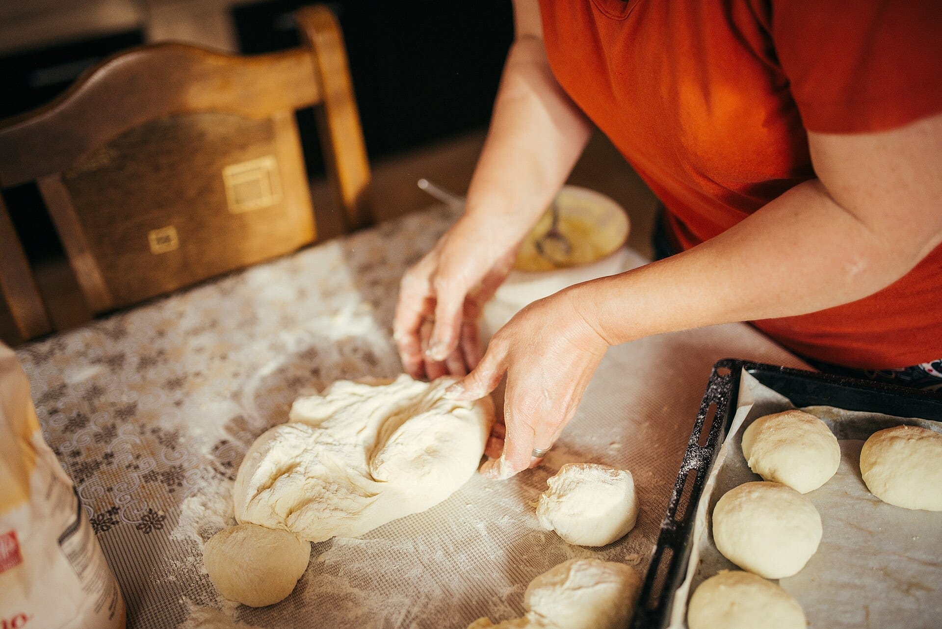 Floury hands shaping dough on a kitchen table beside a tray of rounded dough pieces